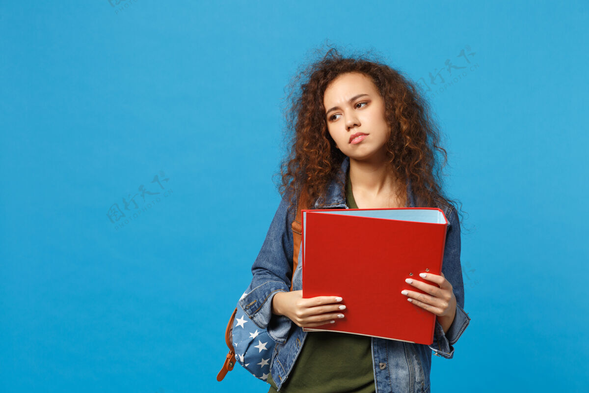 包年轻的非洲裔美国女孩十几岁的学生在牛仔衣服背包举行文件夹隔离在蓝色墙上纸复制空间文件