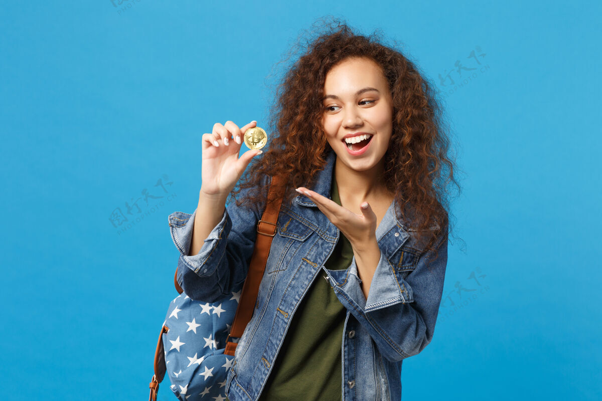 货币年轻的非裔美国少女 穿着牛仔服 背包里装着比特币 孤零零地站在蓝色的墙上学生现金牛仔裤
