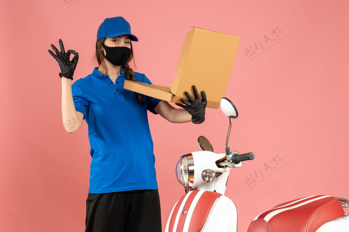 盒子戴着医用口罩手套的年轻快递女孩站在摩托车开箱旁 在柔和的桃色背景下做着戴眼镜的手势手势医学立场