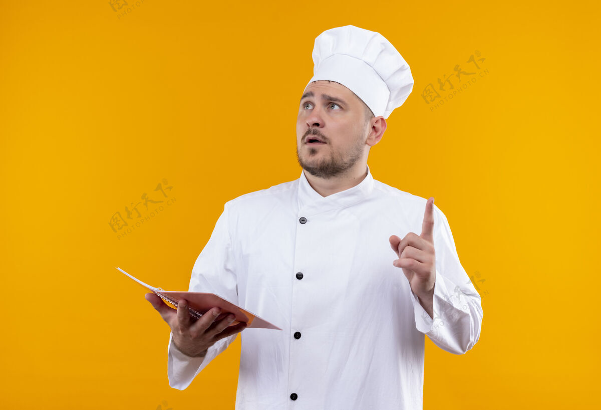 手指穿着厨师制服的年轻帅哥厨师手拿记事本 抬起手指孤立地看着橙色的墙上提高年轻垫