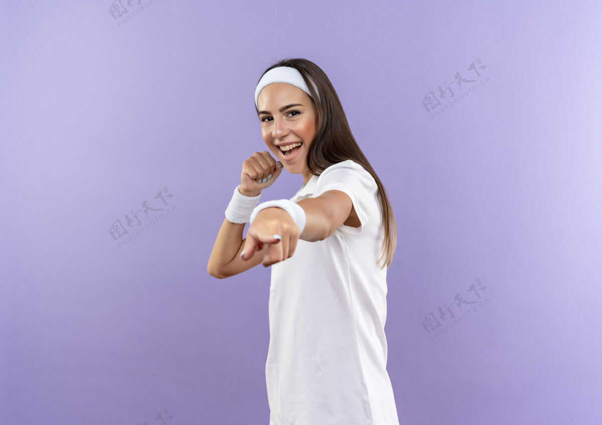 快乐戴着头带和腕带的快乐漂亮的运动女孩举起拳头 孤立地指着紫色的墙上漂亮头带拳头