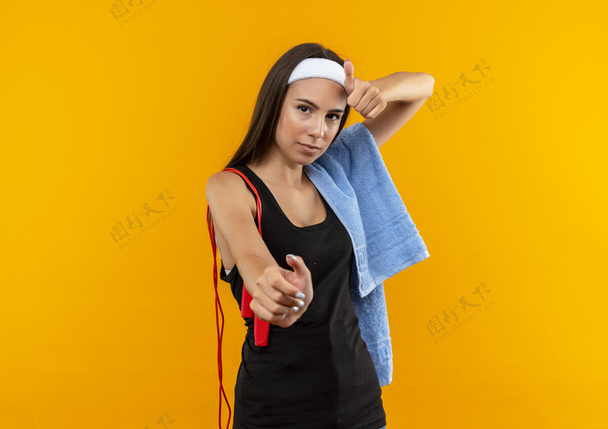 漂亮自信的年轻漂亮的运动女孩戴着头带和腕带 肩上有毛巾和跳绳 伸出手 在橘色的墙上竖起大拇指展示年轻头带