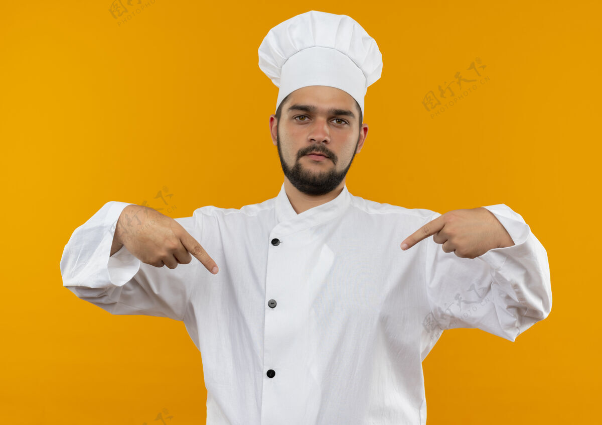 男性身着厨师制服的自信的年轻男厨师指着隔离在橙色墙上的自己指点自信制服