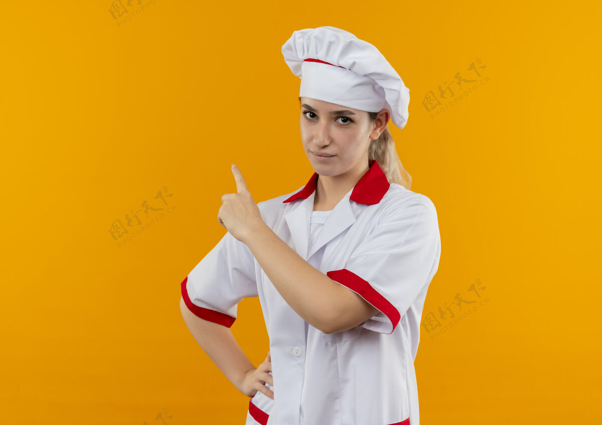 腰自信的年轻漂亮厨师身着厨师制服 手放在腰上 孤立地指着橙色的墙上烹饪年轻制服