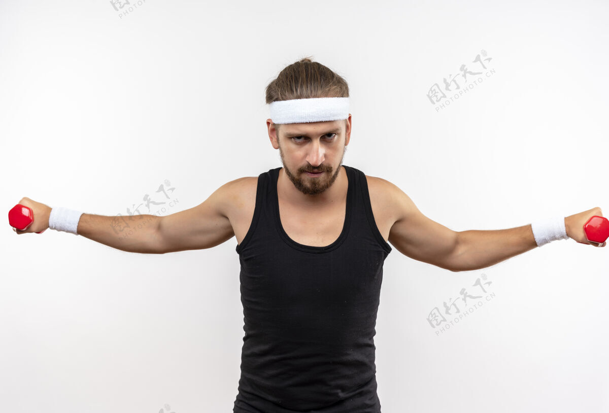 自信自信的年轻帅气的运动型男人戴着头带和腕带举着哑铃伸开双臂隔离在白色的墙上运动年轻开放