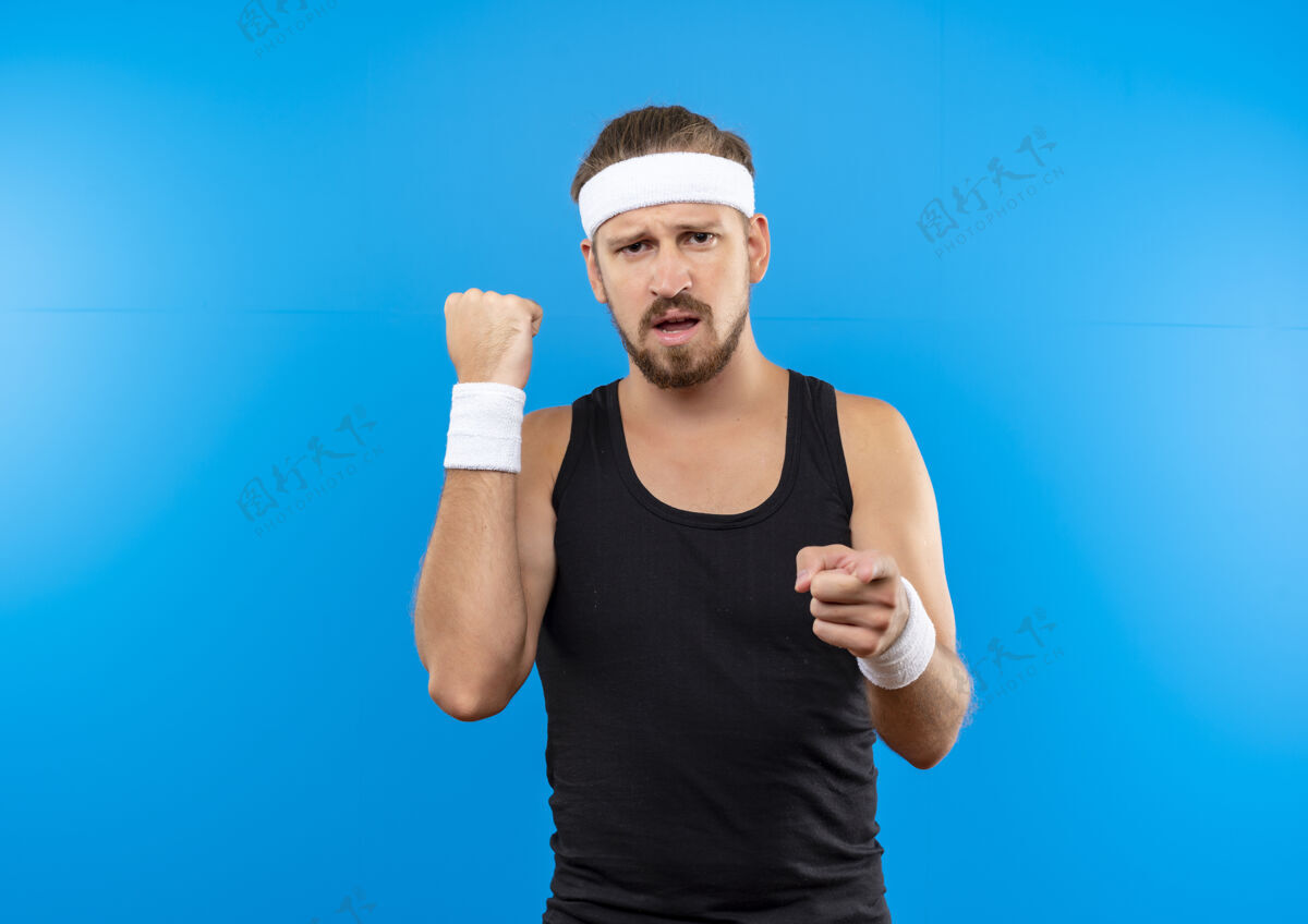 年轻自信的年轻帅气的运动型男人戴着头带和腕带握紧拳头 孤立地指着蓝色的墙壁和复制空间尖拳头自信