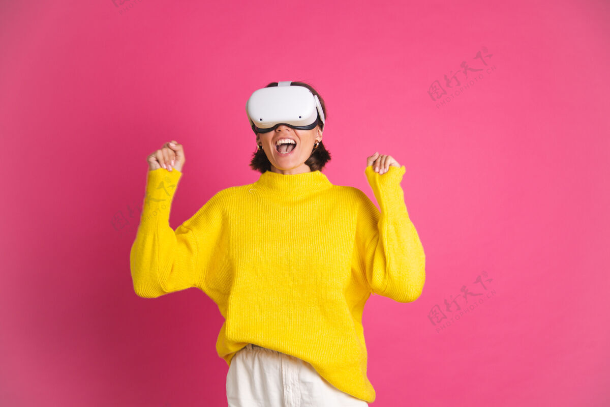 创新穿着亮黄色毛衣的美女戴着粉色的虚拟现实眼镜高兴地跳起来握紧拳头赢家的手势青少年视频展示