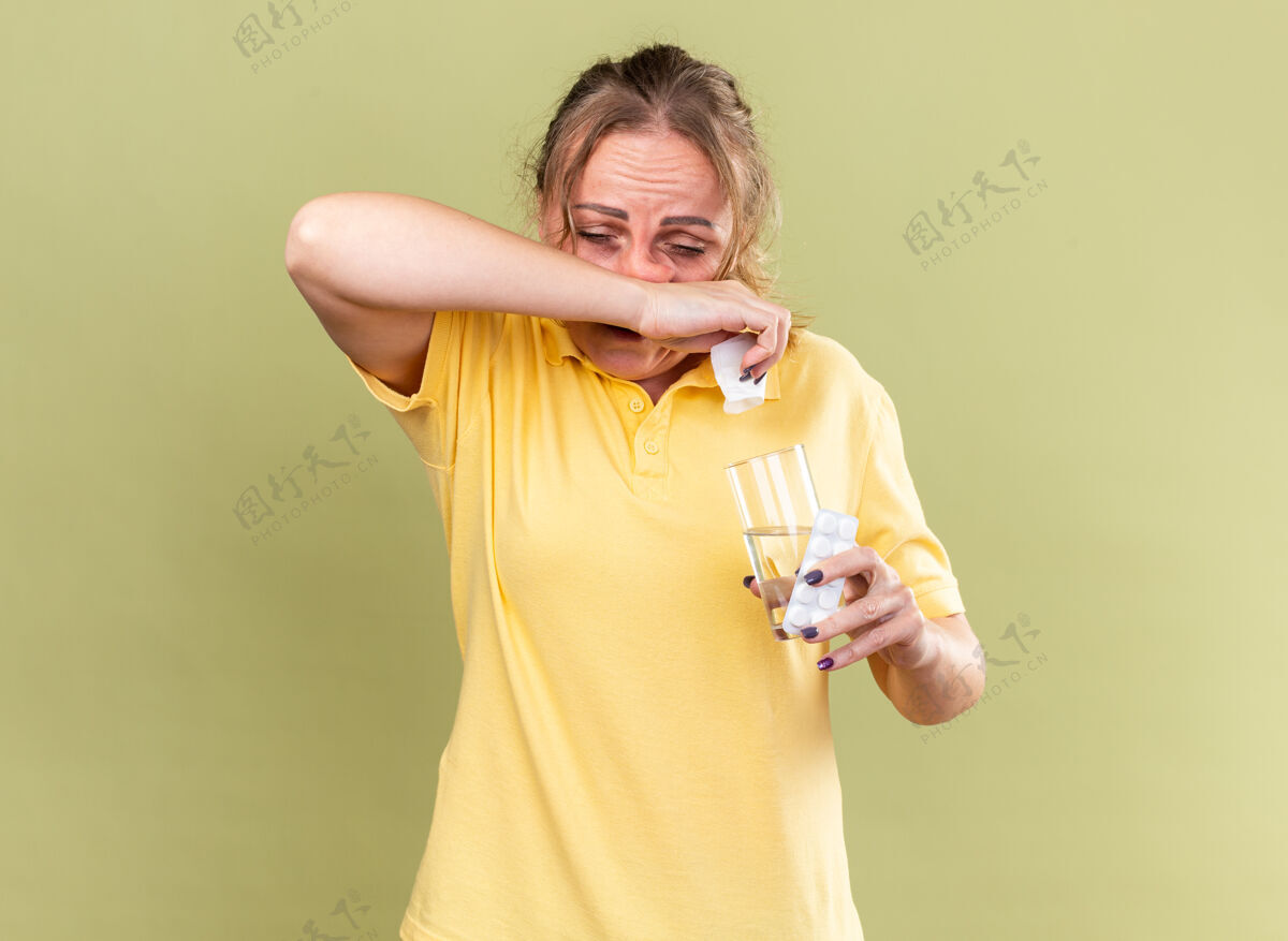 抱着一个穿着黄色衬衫的不健康的女人站在绿色的墙壁上 手里拿着一杯水和药片擦着流鼻涕 感觉很糟糕站着不健康药片