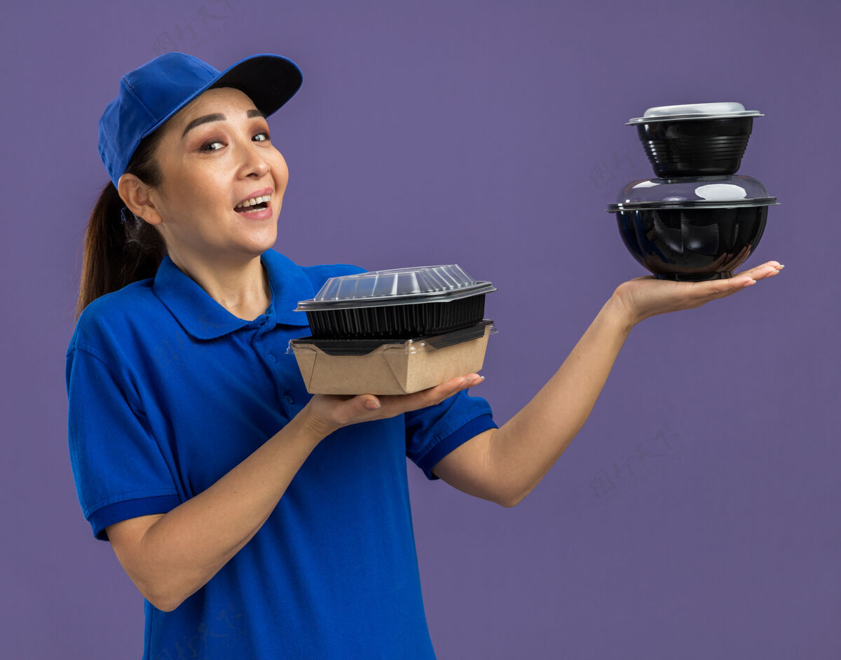 女人身穿蓝色制服 头戴鸭舌帽 拿着食品包的年轻送货员站在紫色的墙上 快乐而欢快地微笑着欢呼年轻帽子