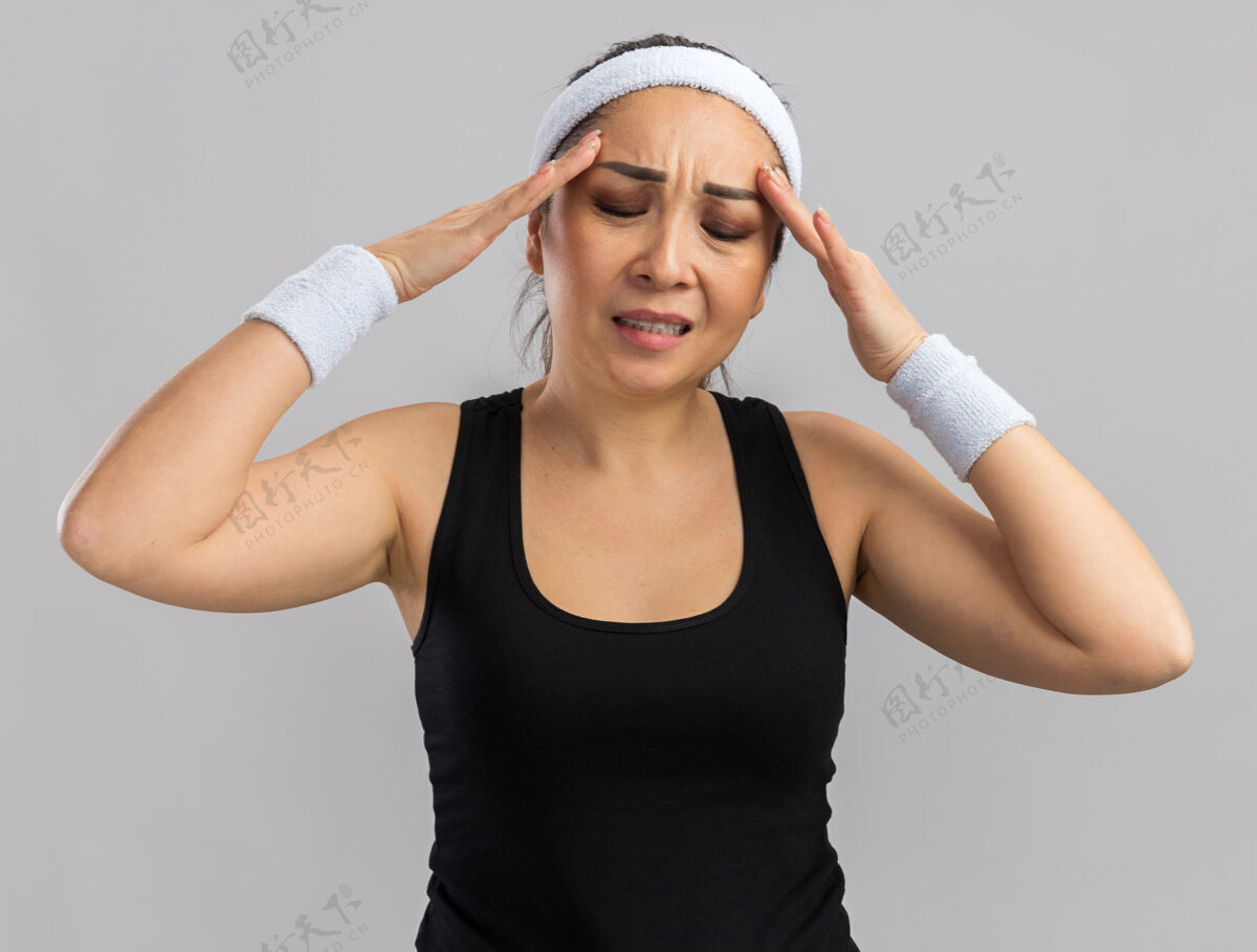头戴着头带和袖带的年轻健身女士站在白墙上 头上摸着头 看起来很不舒服 头痛抚摸年轻站立