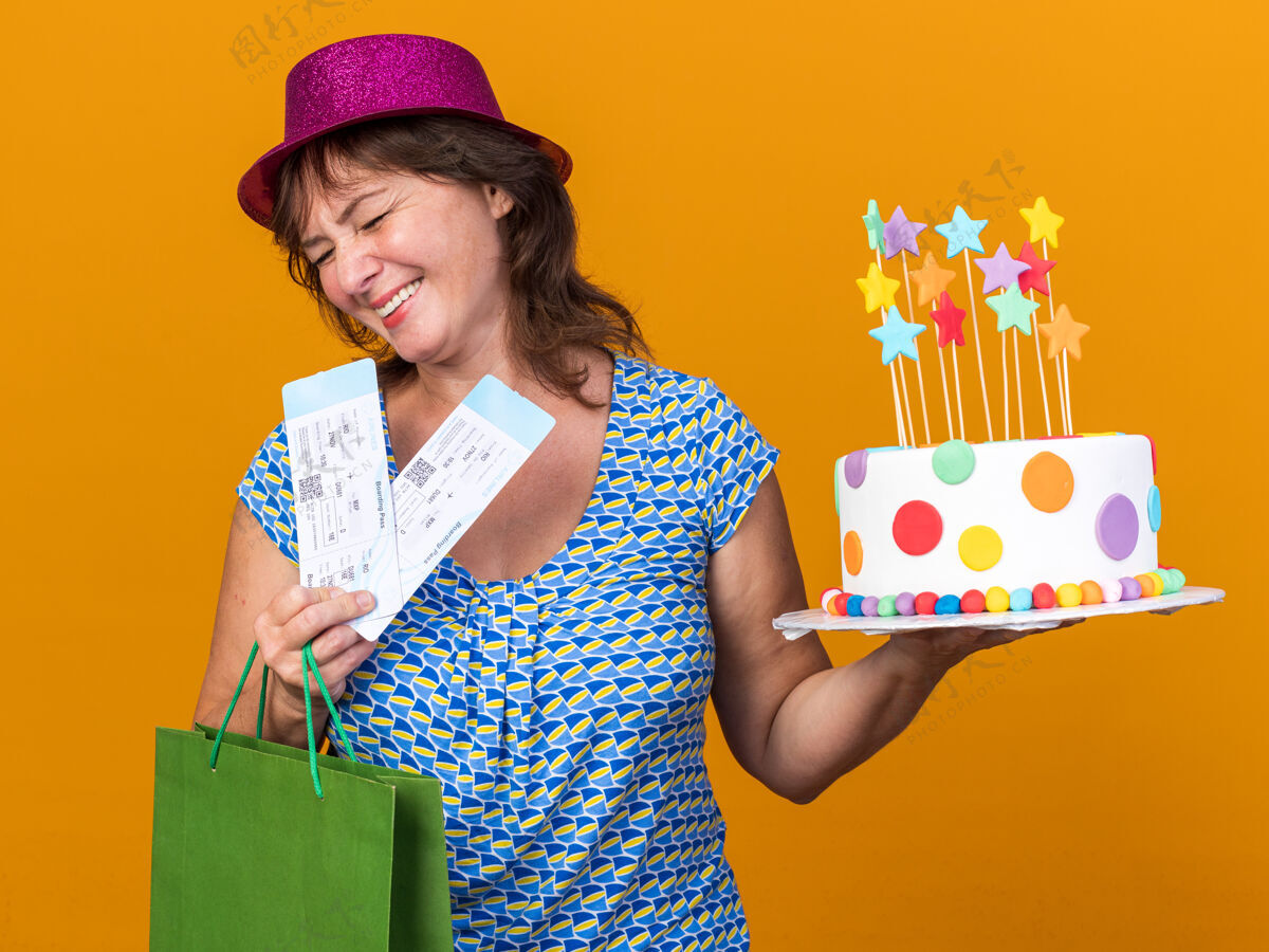 蛋糕中年妇女戴着派对帽 手里拿着带礼物的纸袋 手里拿着生日蛋糕和机票 开心地微笑着 站在橘色的墙上欢庆生日派对中间女人举行