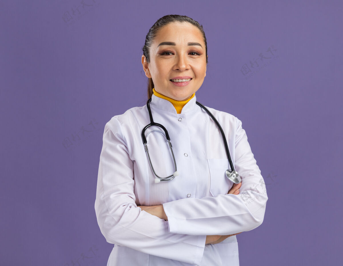 听诊器身穿白色药衣 脖子上戴着听诊器的年轻女医生自信地微笑着双臂交叉着站在紫色的墙上站年轻人女人