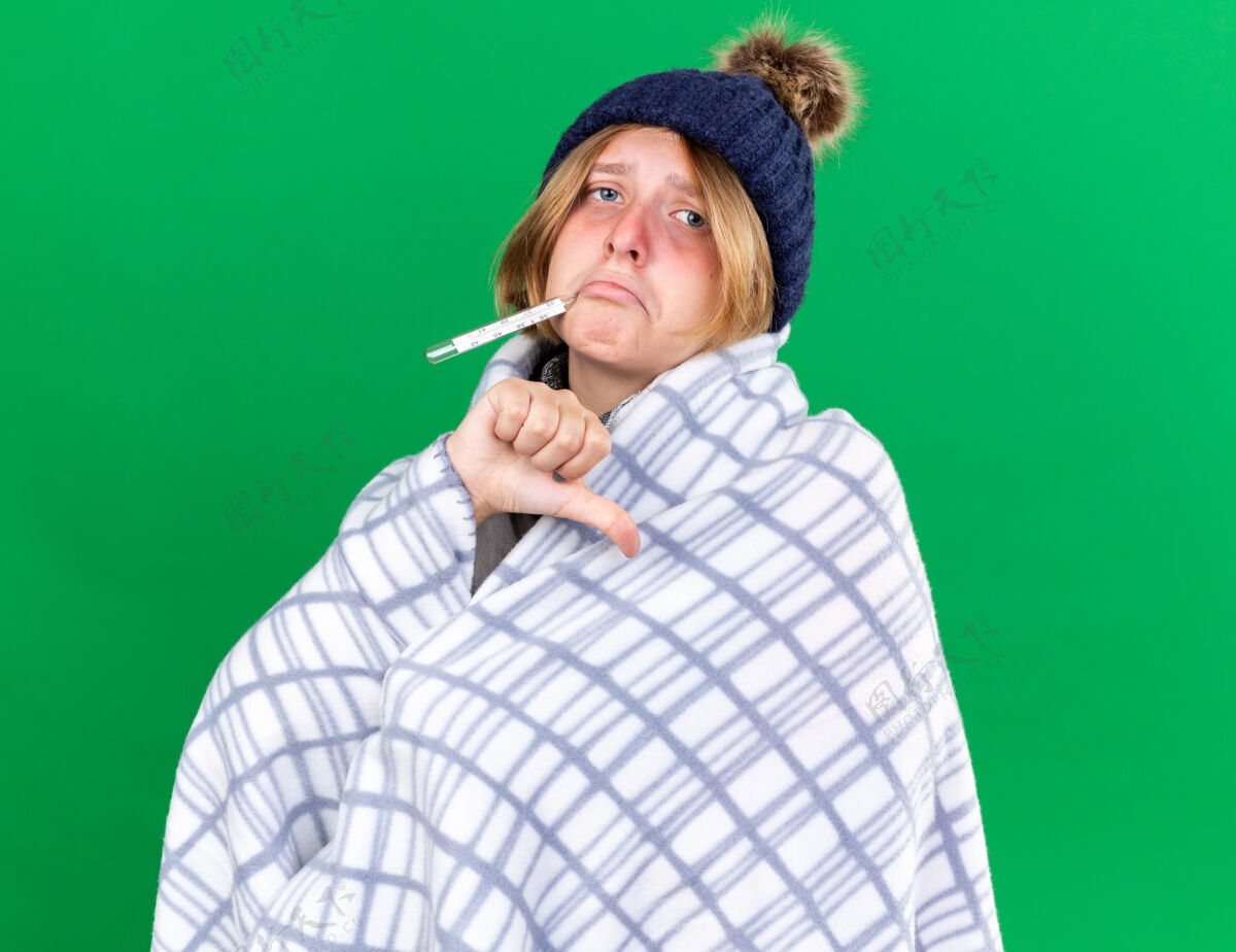拇指不健康的年轻女子裹着毛毯戴着帽子用体温计测量体温患流感发烧大拇指朝下站在绿色的墙上身体不健康发烧