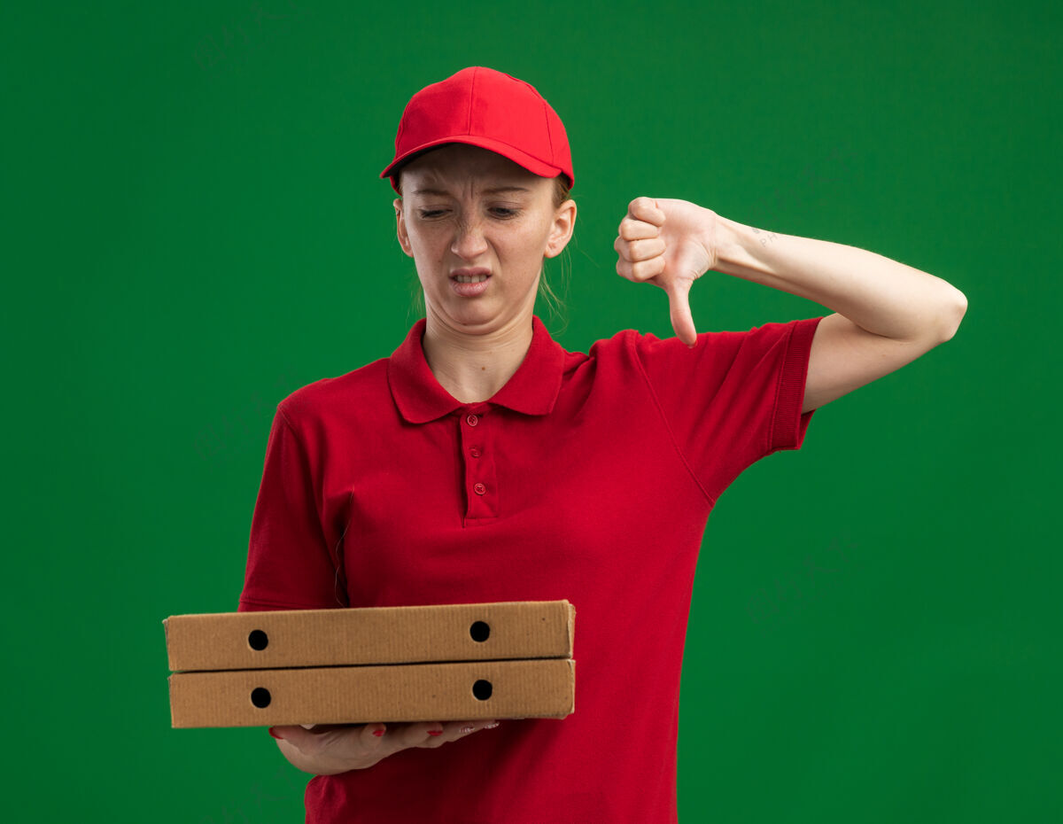 女孩身穿红色制服 头戴鸭舌帽的年轻送货员站在绿色的墙上 不高兴地竖起大拇指看着比萨饼盒不高兴站着年轻
