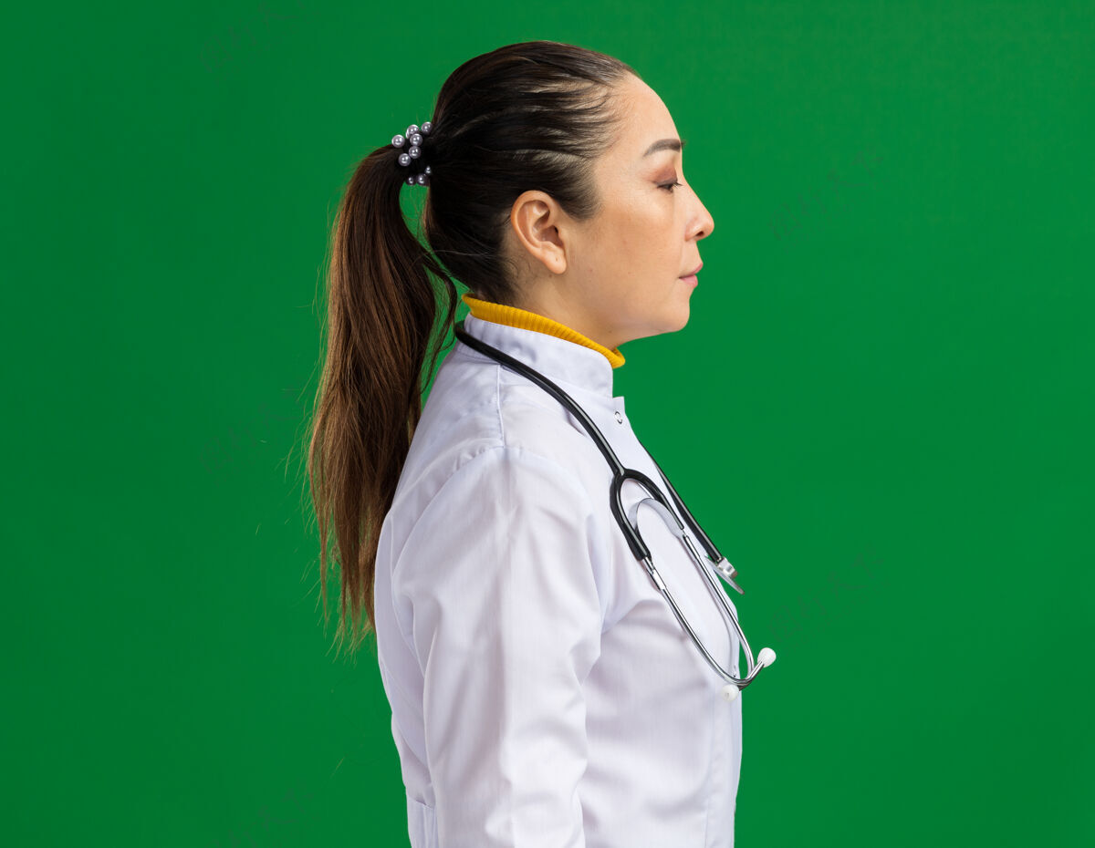 外套年轻的女医生 穿着白药衣 脖子上戴着听诊器 脸贴着绿墙 侧身站着医生听诊器药