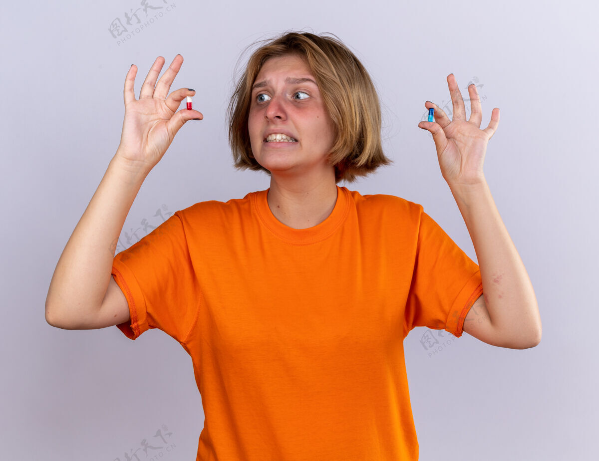 不健康不健康的年轻女子穿着橙色t恤 感觉身体不适 手上拿着感冒药 站在白色的墙上显得困惑和担心女人吃药迷茫