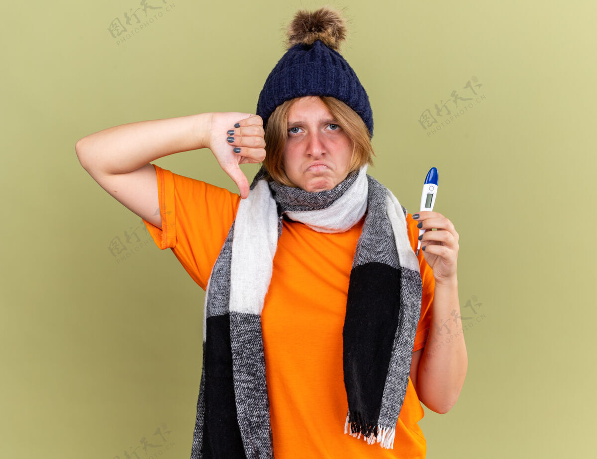 发烧不健康的年轻女子戴着帽子 脖子上围着围巾 感觉不舒服 拿着数字体温计 患流感发烧 站在绿色的墙壁上大拇指朝下感觉数码显示