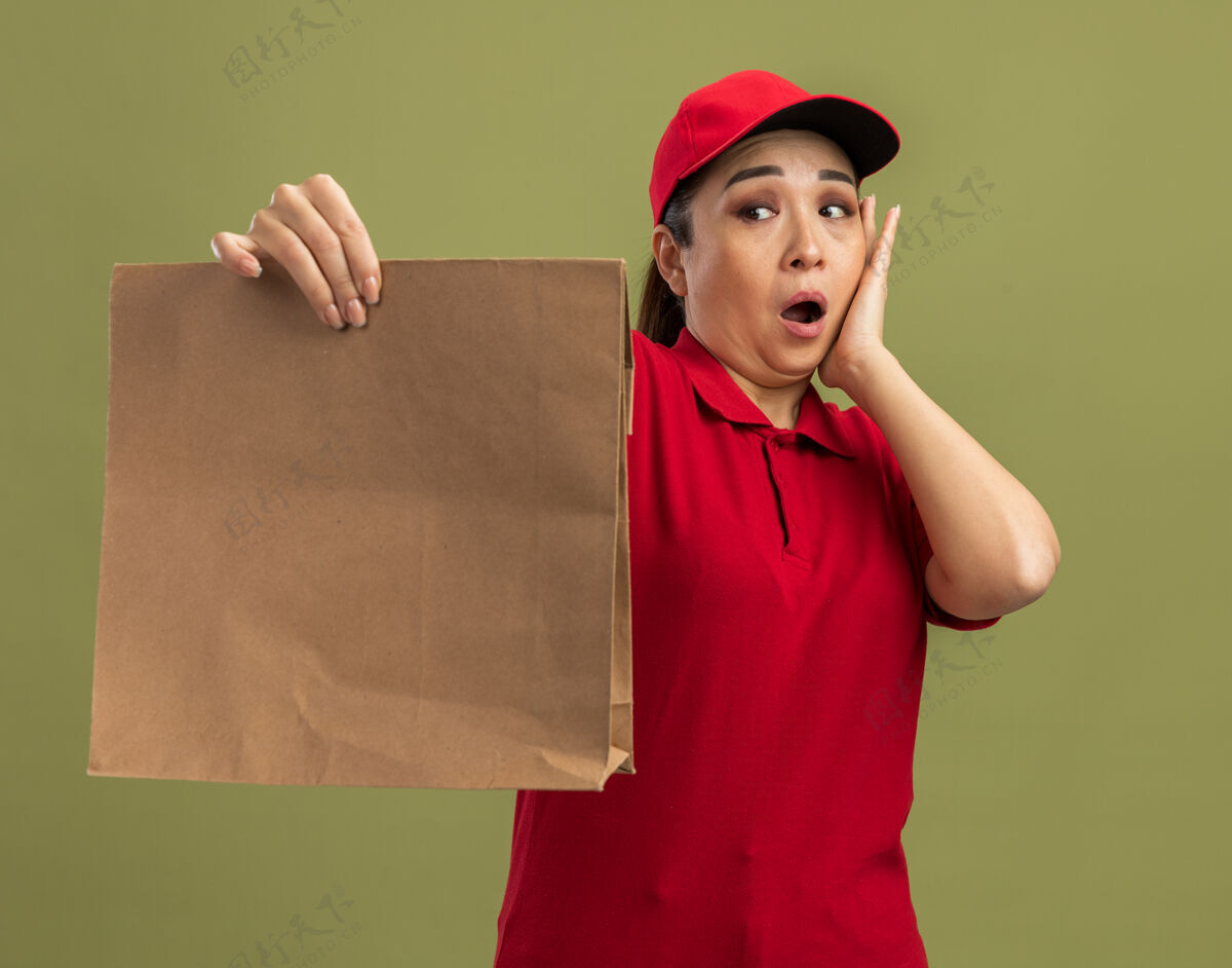 送货年轻的送货员身穿红色制服 戴着帽子 手里拿着纸包 站在绿色的墙边困惑而惊讶地看着它迷茫帽子制服