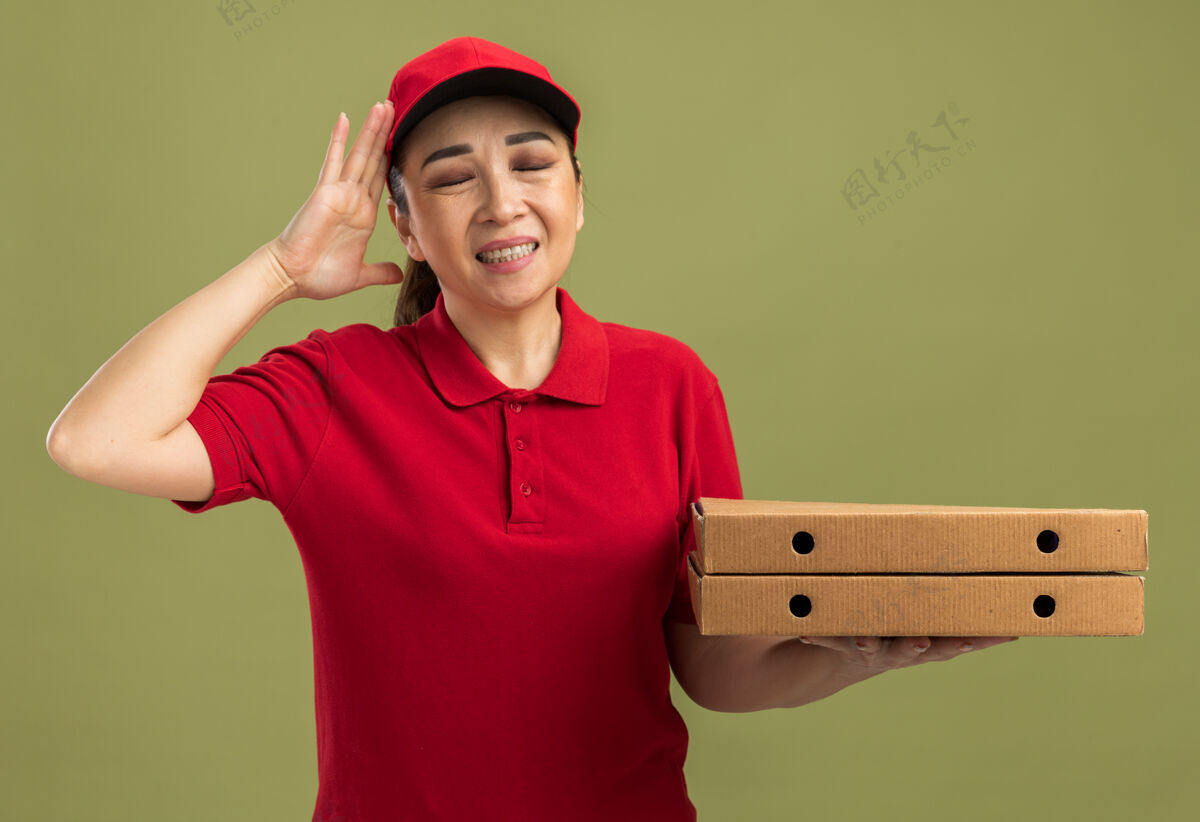 胳膊年轻的送货员身穿红色制服 头戴鸭舌帽 手里拿着披萨盒 站在绿色的墙上 看起来既恼火又恼火盒子披萨年轻人