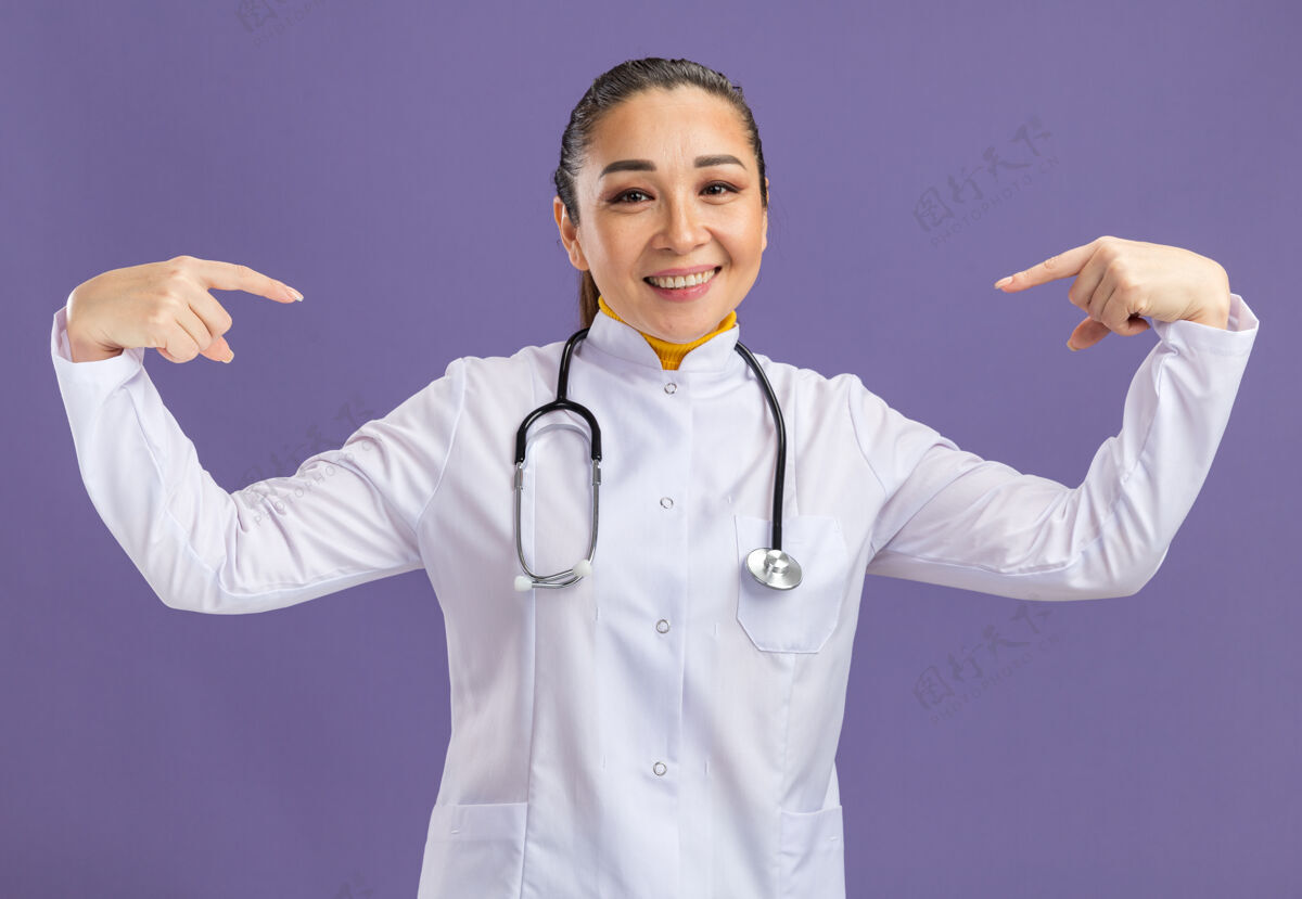 自信身穿白色药衣 脖子上戴着听诊器的年轻女医生自信地微笑着指着站在紫色墙上的自己听诊器微笑医生