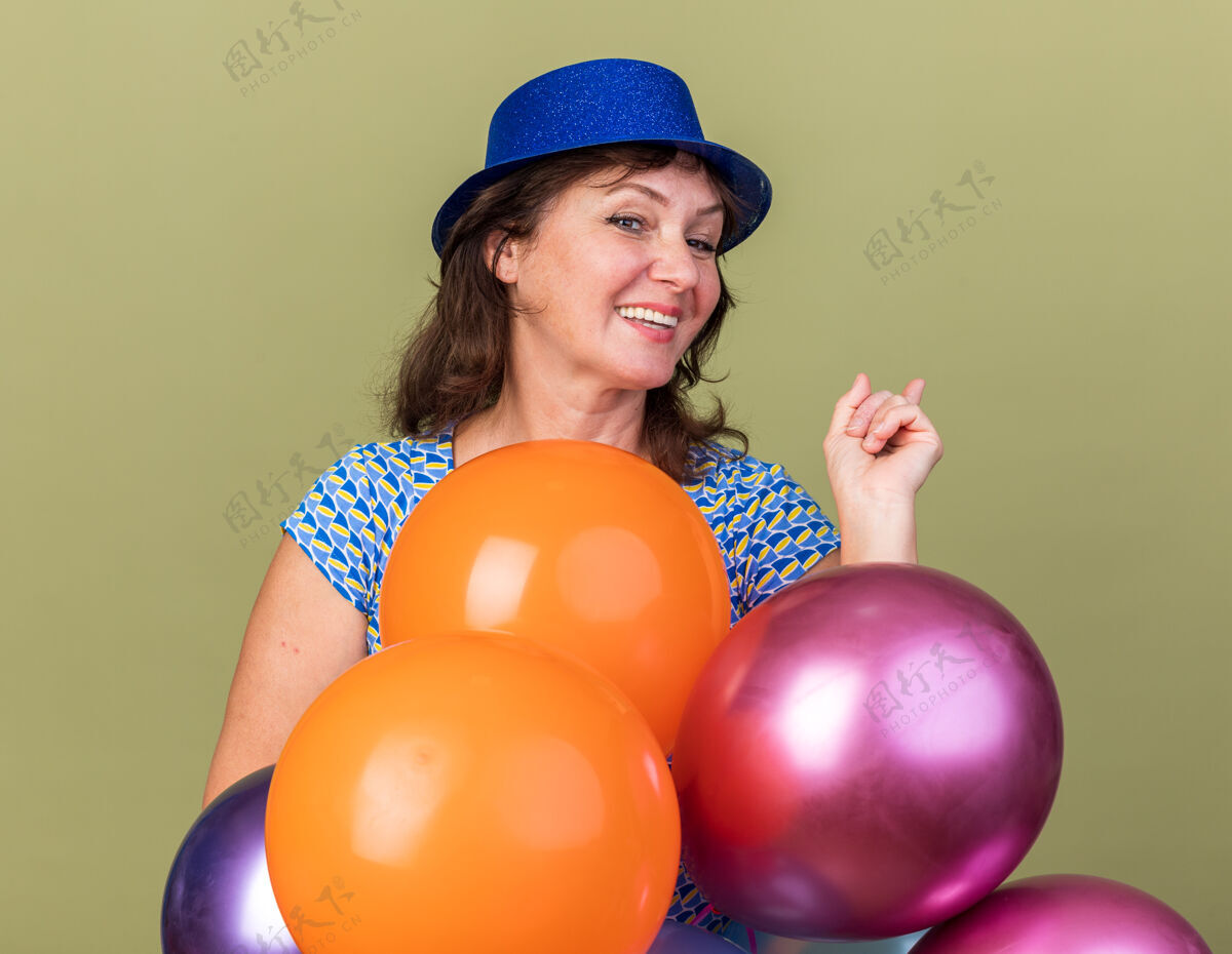 气球快乐快乐的中年妇女戴着派对帽 手持一束五颜六色的气球 微笑着站在绿色的墙上庆祝生日派对年龄微笑欢呼