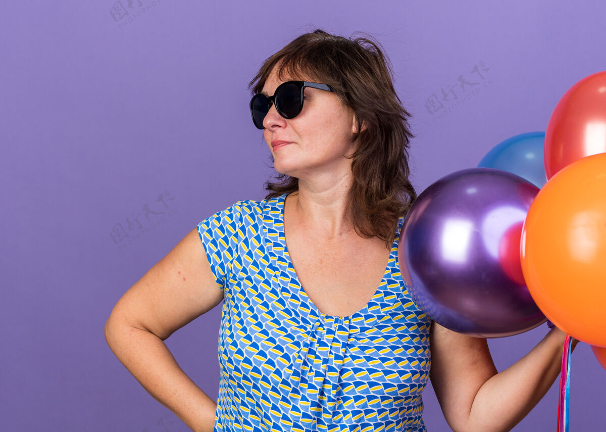 女人中年妇女戴着眼镜 手里拿着一堆五颜六色的气球 表情严肃地站在紫色的墙上庆祝生日派对中间眼镜脸