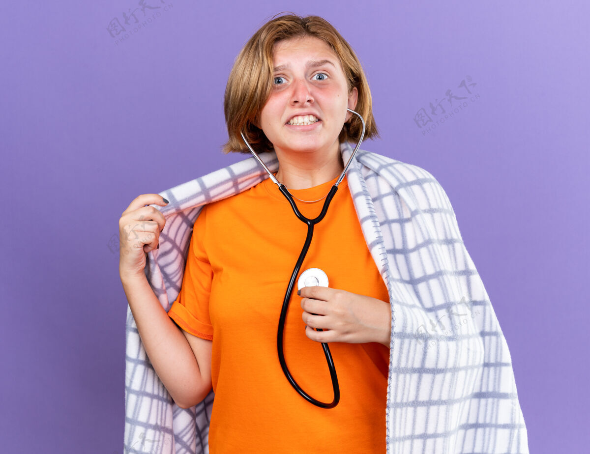 包装不健康的年轻女子裹着温暖的毯子 感觉不舒服 站在紫色的墙上 用听诊器听着自己的心跳 看起来很担心不健康女人感觉