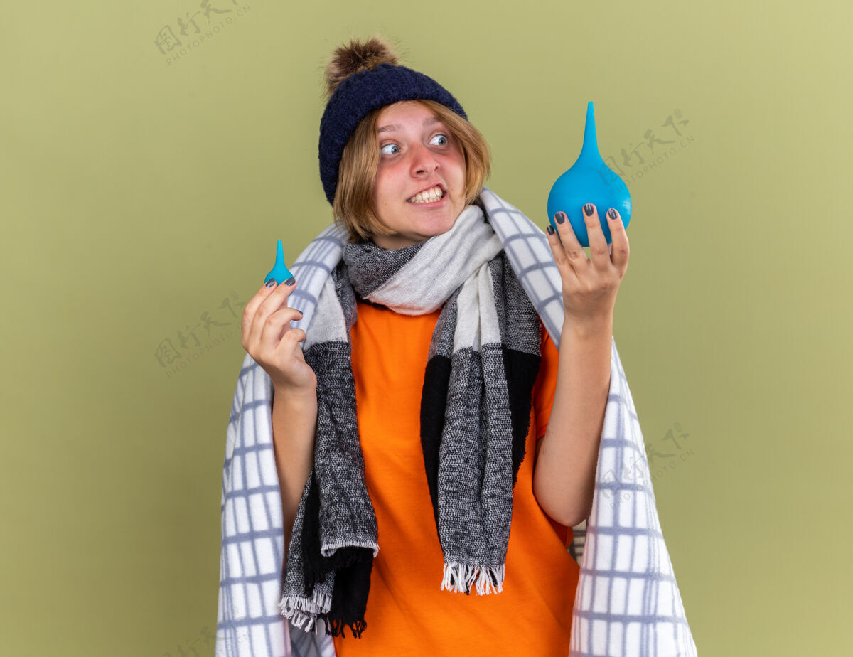 帽子不健康的年轻女子裹着毯子戴着帽子和围巾拿着灌肠剂困惑地站在绿色的墙壁上感觉不舒服不健康感觉围巾