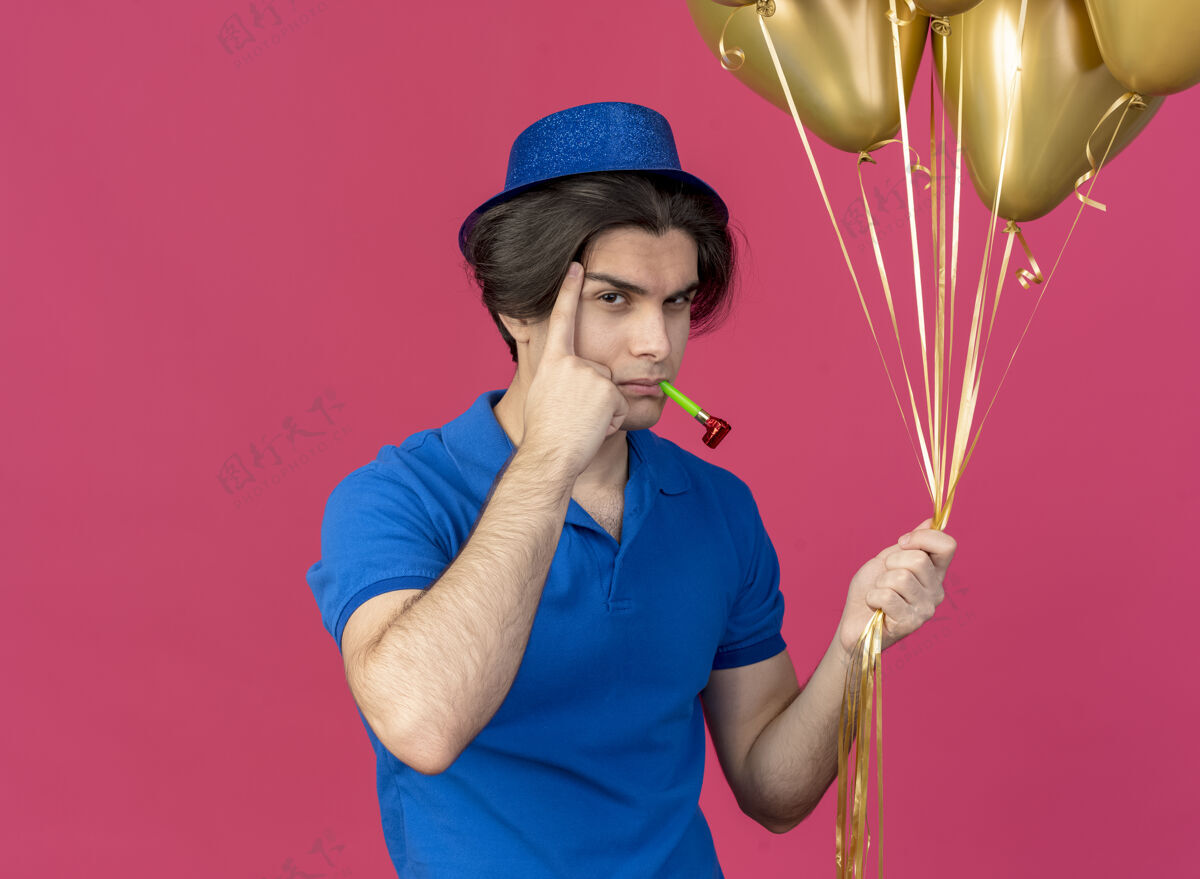 帅气自信英俊的白人男子戴着蓝色的派对帽 手指放在太阳穴上 手持氦气球吹派对哨子氦拿男人