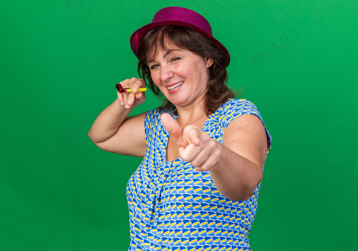 手指戴着派对帽的中年妇女手持哨子 食指微笑 站在绿色的墙上欢庆生日派对目录生日年龄