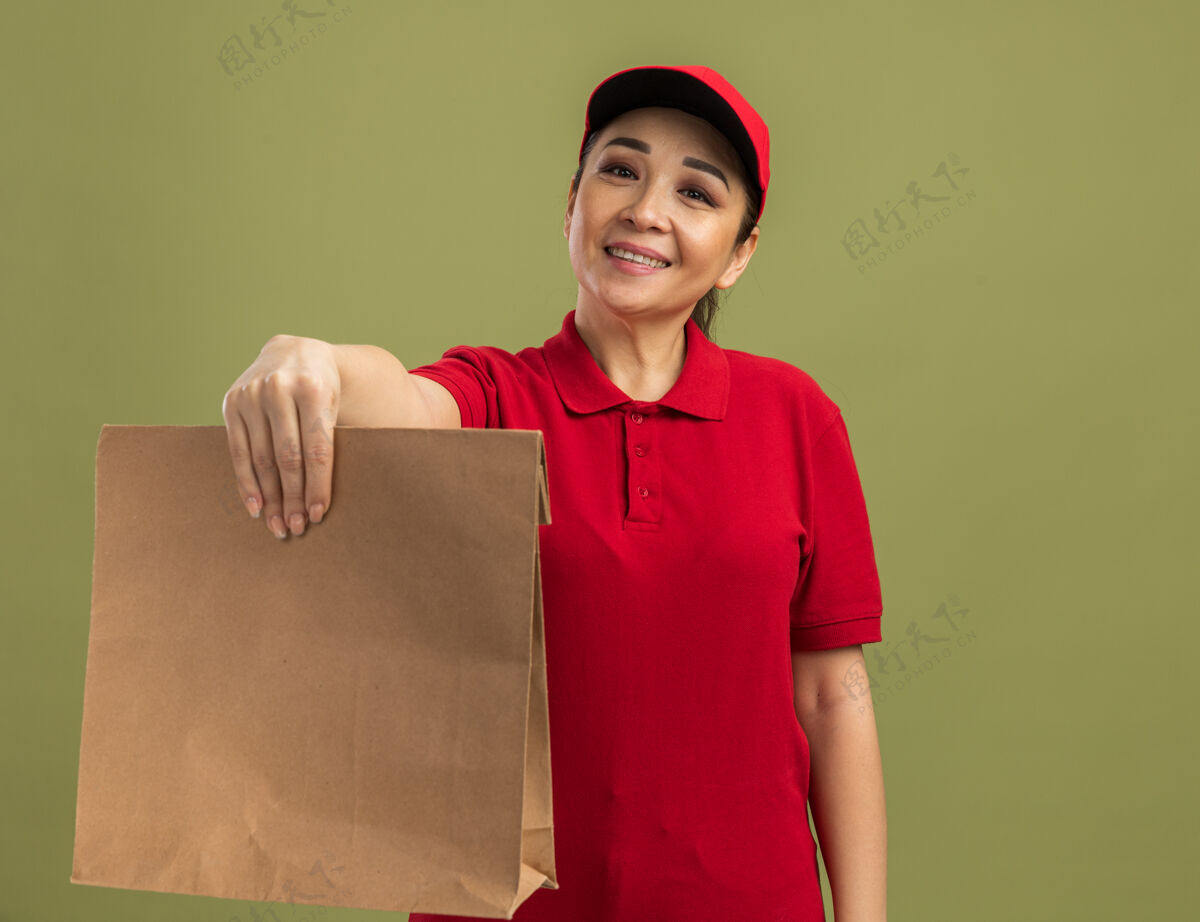 脸身穿红色制服 头戴鸭舌帽 面带微笑的年轻送货员站在绿色的墙边 快乐而积极持有积极女人