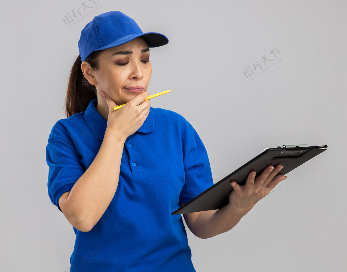 年轻身穿蓝色制服 戴着帽子 拿着写字板和钢笔的年轻女送货员站在白墙上沉思着女人表情制服