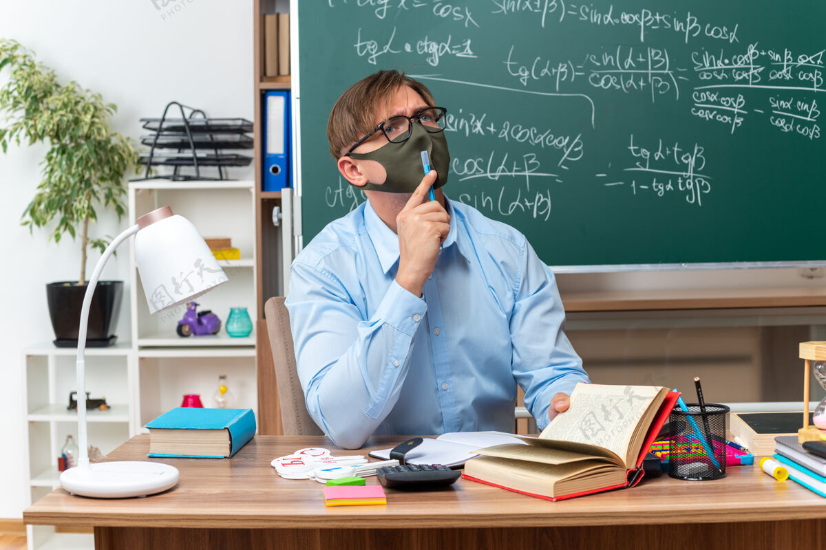 男年轻的男老师戴着眼镜 戴着口罩 困惑地抬头望着 坐在课桌旁 教室的黑板前放着书和笔记保护黑板拼图