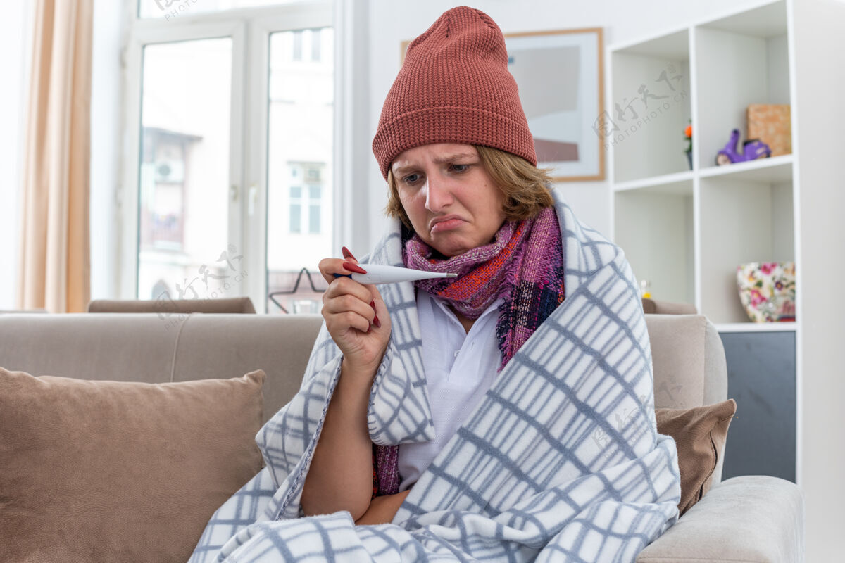 围巾不健康的年轻女子戴着帽子 脖子上围着暖和的围巾 拿着温度计检查体温 感觉不舒服 患了感冒和流感 坐在明亮的客厅里的沙发上看起来很担心检查房间沙发