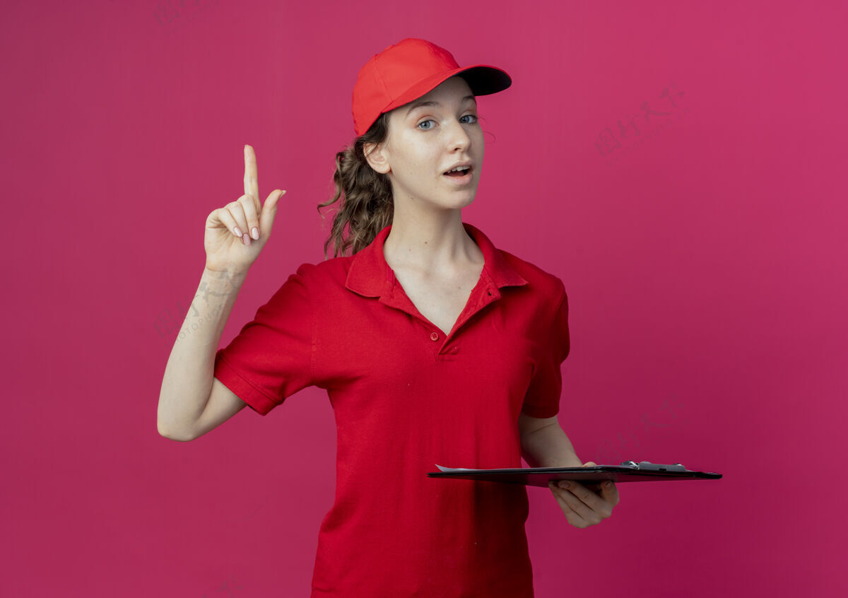 提高印象深刻的年轻漂亮的送货女孩在红色制服和帽子举行剪贴板和提高手指在深红色的空间红色漂亮帽子