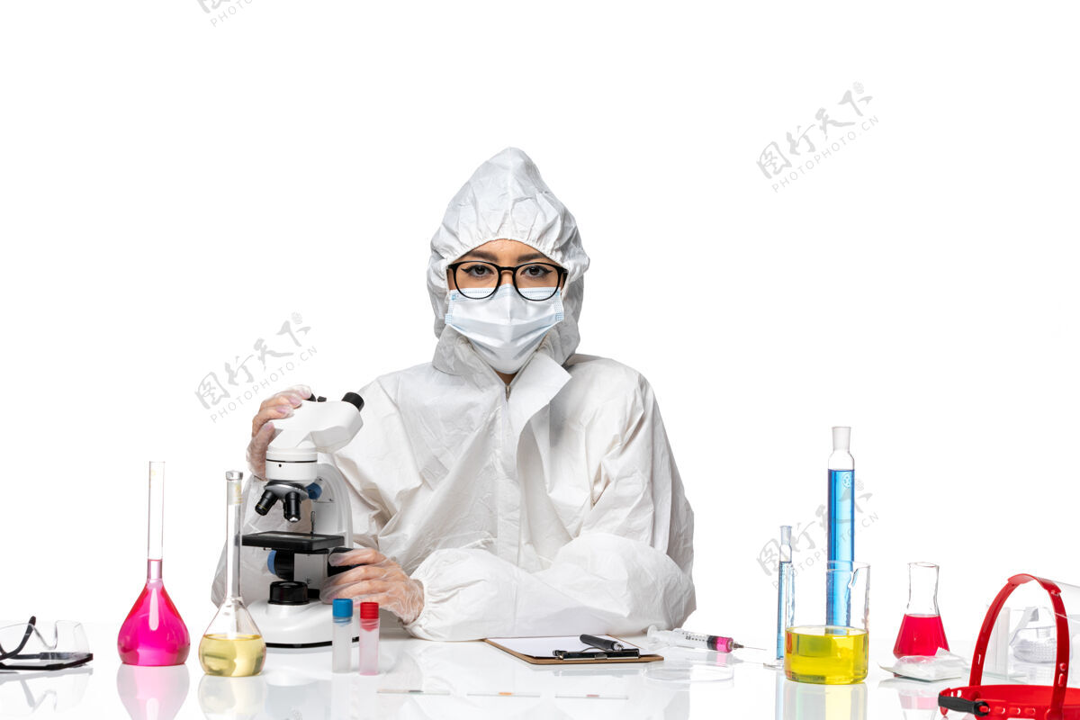使用正面图身着特殊防护服的年轻女化学家在白色办公桌上使用显微镜病毒实验室共价健康化学面具显微镜化学