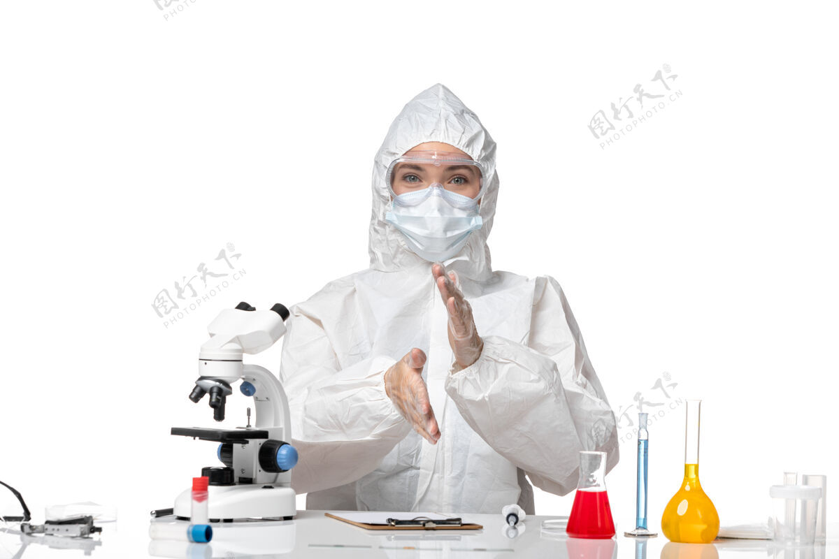 西装正面图年轻女医生穿着防护服戴着口罩 由于科维德坐在白色背景上微笑病毒大流行飞溅科维德-医院医学大流行