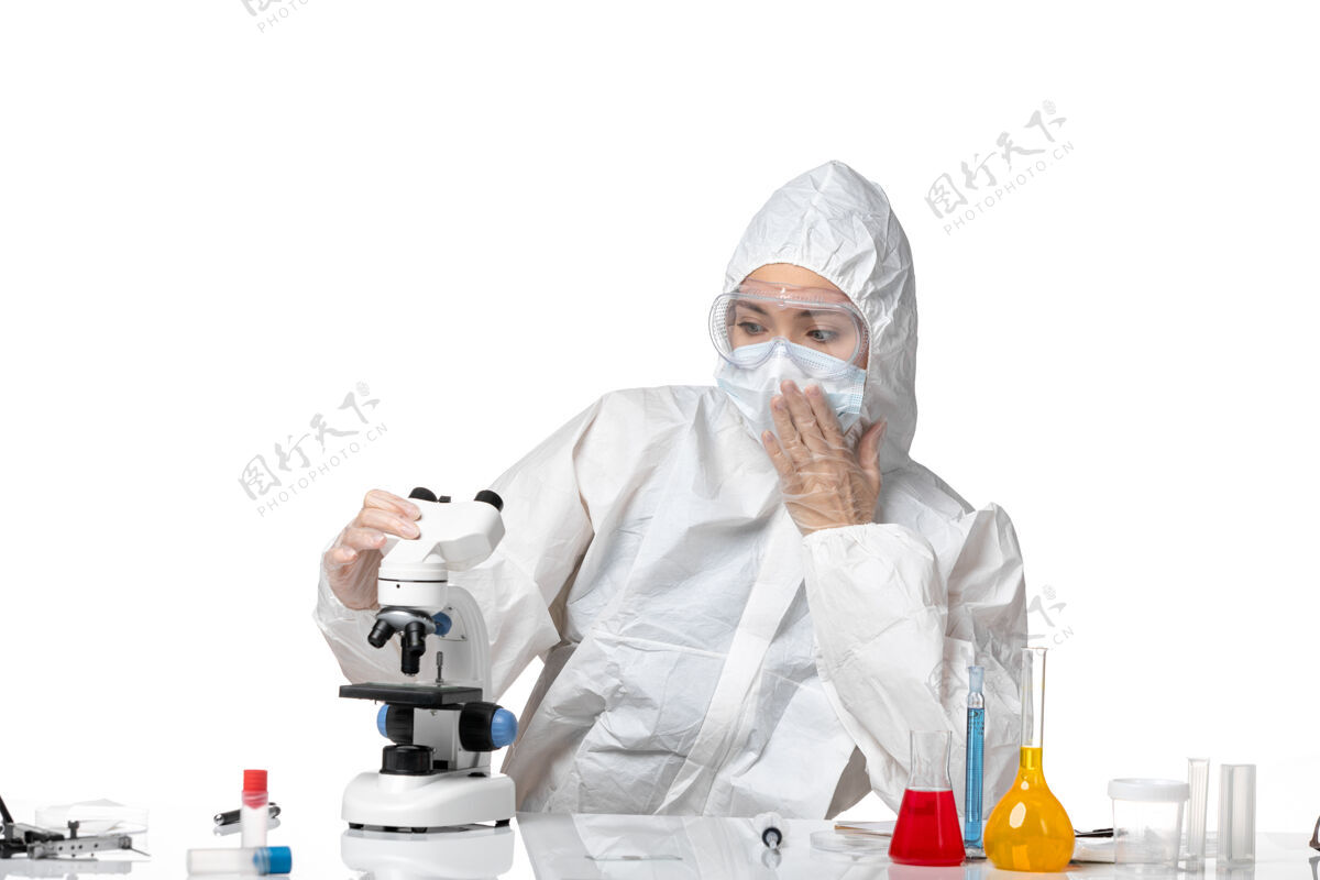 药品正面图年轻女医生穿着白色防护服 戴着口罩 由于使用显微镜在白色办公桌上大流行 飞溅病毒健康使用外套医生