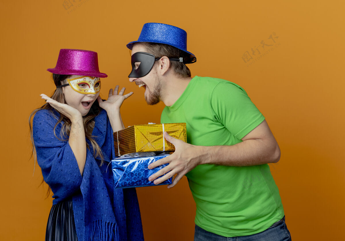 帅气快乐的帅哥戴着蓝色帽子戴着化装眼罩拿着礼盒看着惊讶的年轻女孩戴着粉色帽子和化装眼罩举手看着礼盒举行惊喜年轻