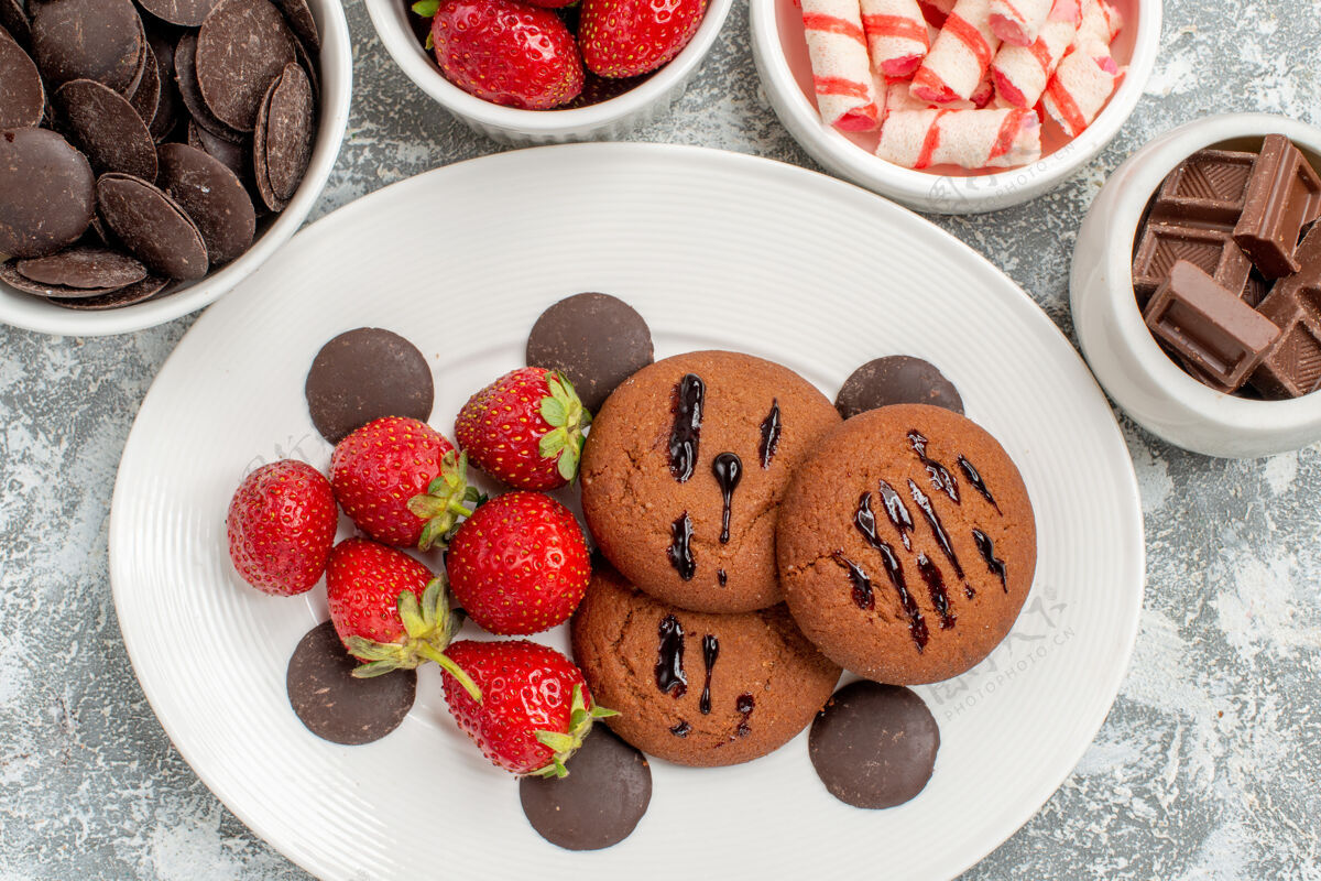 盘子特写镜头：白色椭圆形盘子上的饼干草莓和圆形巧克力 灰白色的桌子上有糖果草莓和巧克力早餐食物浆果