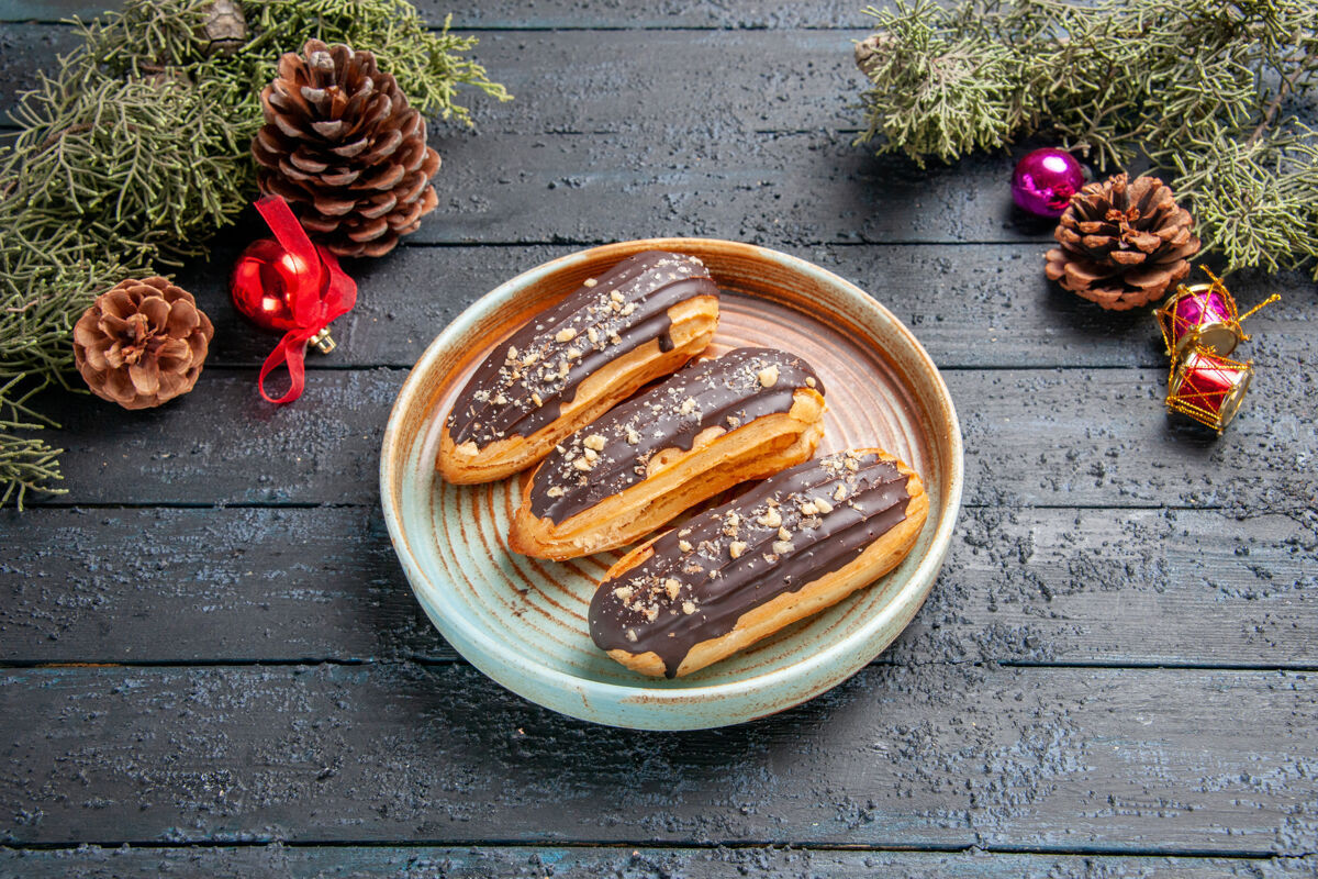黑暗底部视图巧克力eclairs上的椭圆形板松果圣诞玩具冷杉树叶上的黑色木制背景木头椒盐卷饼食物