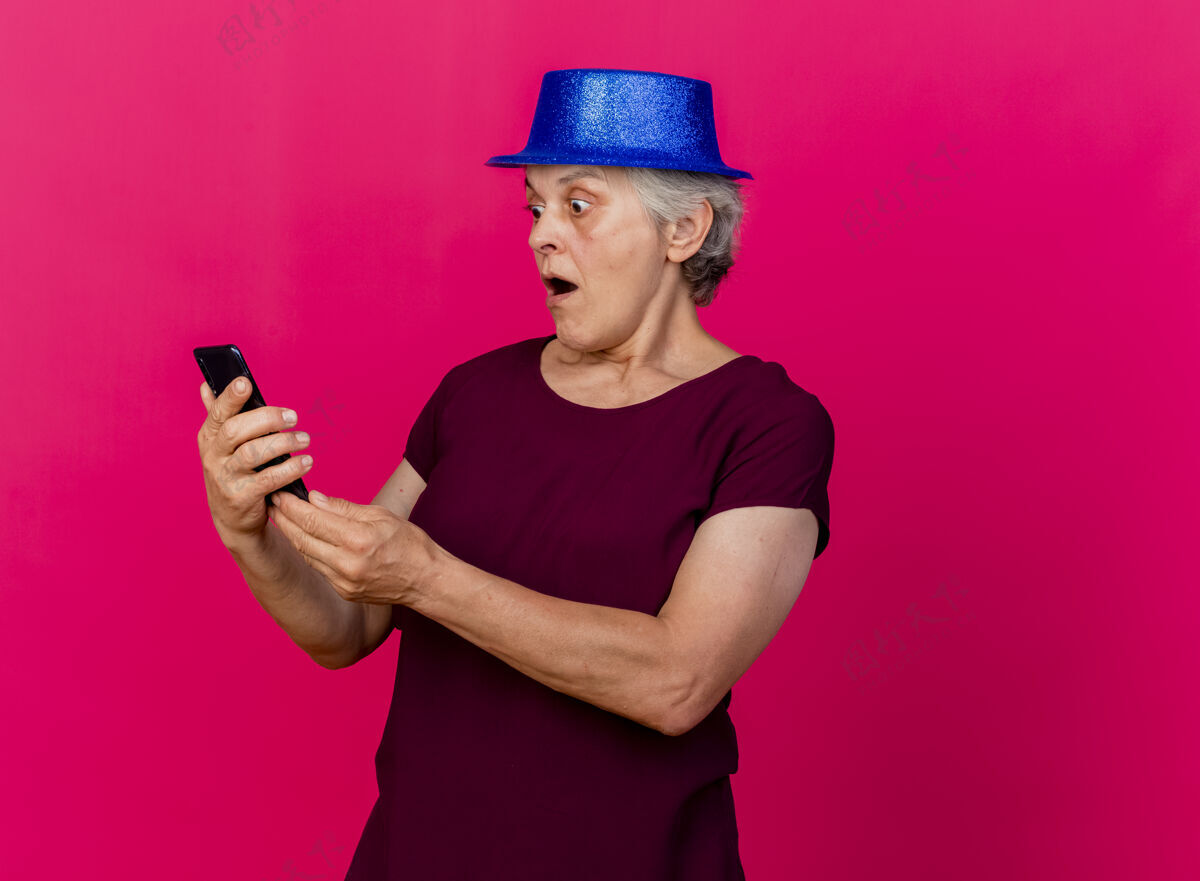 女人惊讶的老妇人戴着聚会的帽子拿着手机看着粉红色的帽子电话派对