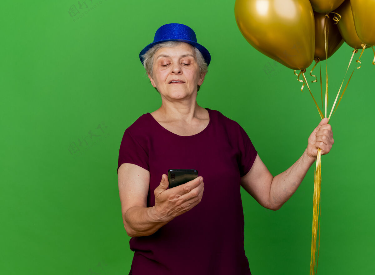 电话戴着派对帽的自信的老妇人手持氦气球 看着绿色的手机气球帽子老人