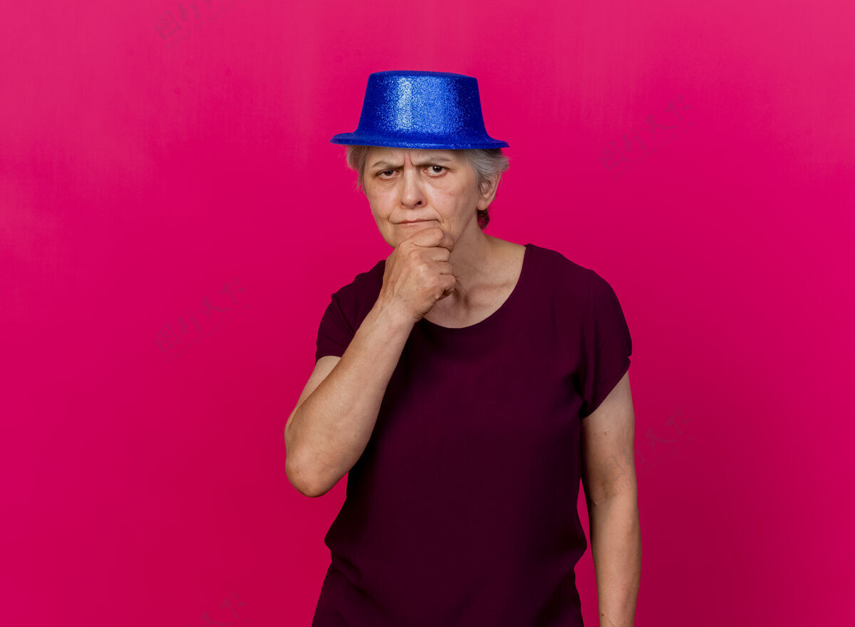 派对困惑的老妇人戴着聚会帽 手放在下巴上看着粉红色的相机老人女人帽子