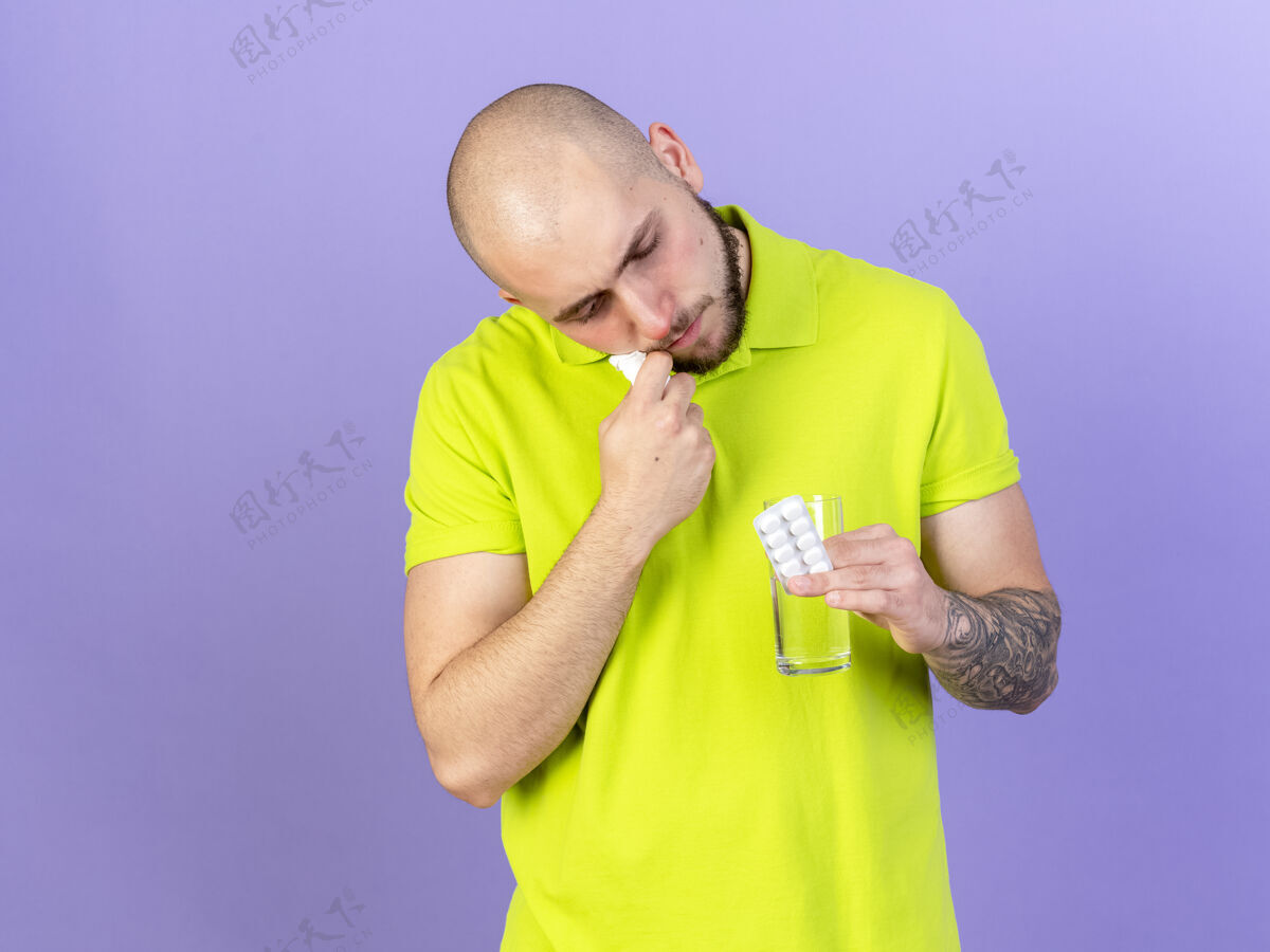 疾病自信的年轻白种人拿着纸巾看着一杯水和一包紫色的药丸男人年轻信心