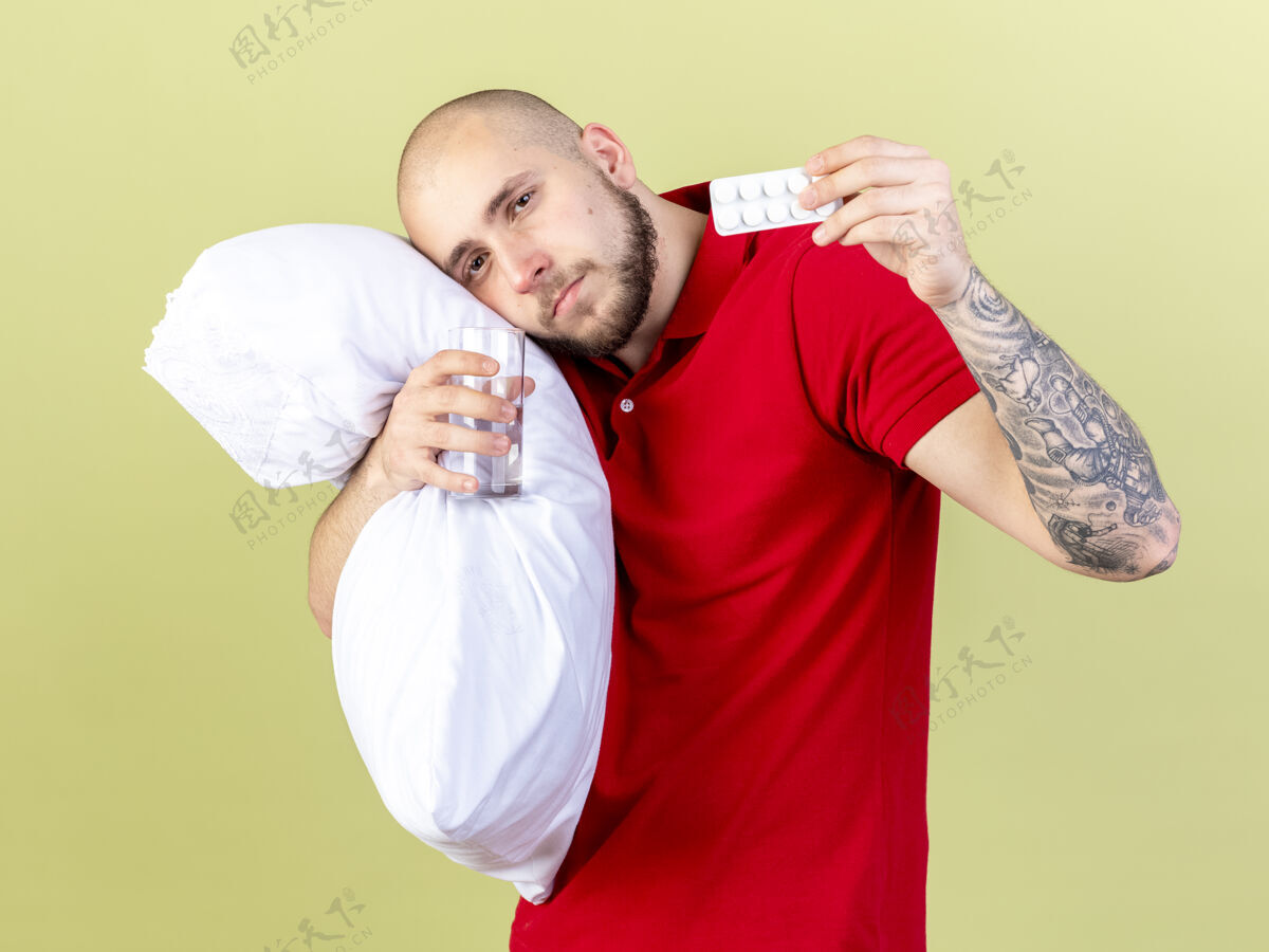 男人高兴的年轻白人病人抱着枕头 抱着一杯水和一包橄榄绿的药丸绿色请医药