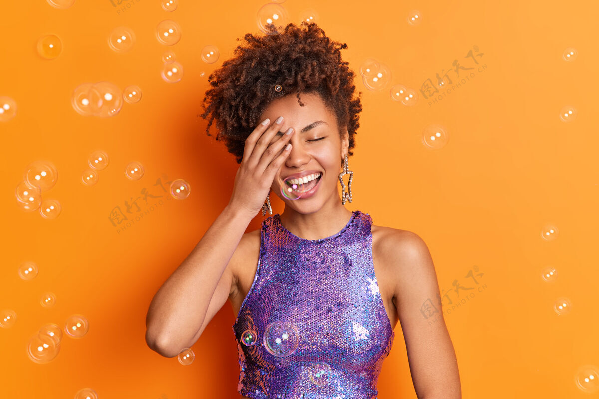 休闲积极的非裔美国妇女用手捂着脸 笑容灿烂 洁白的牙齿 穿着时髦的衣服 隔离在橙色的墙上 周围有肥皂泡大笑肖像室内