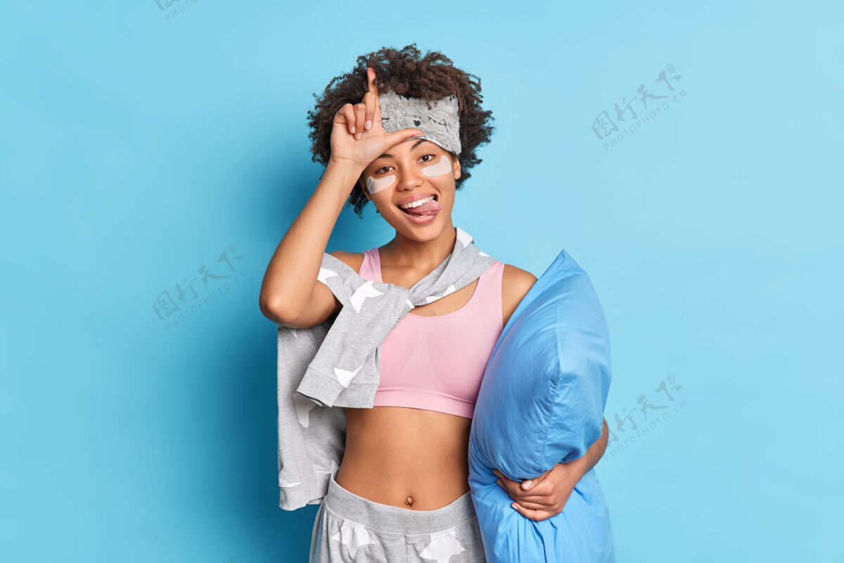 蒙眼有趣的女人做失败者的手势很有趣穿着睡衣拿着柔软的枕头伸出舌头隔着蓝色的墙睡眠失败者睡衣