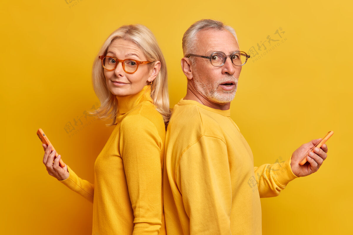 成熟两位上了年纪的男女朋友背对背地站着 戴着眼镜 休闲套头衫 使用现代小玩意进行在线交流 黄色墙壁上隔离着短信互联网退休Omg