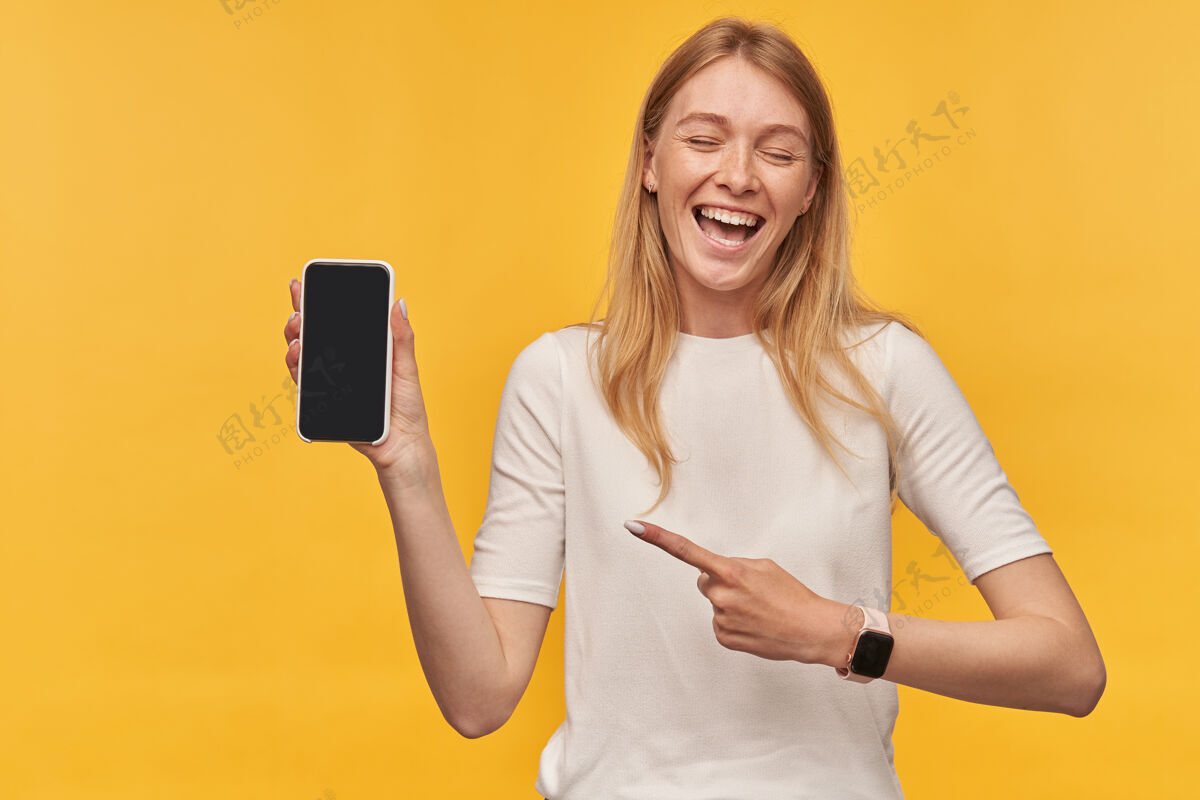 金发快乐美丽的女人 穿着白色T恤 有雀斑 智能手表 笑着指着黄色的黑屏手机女士欢呼请